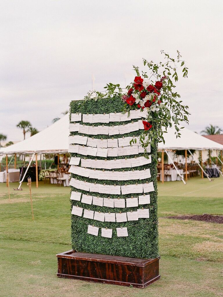 Escort card display on greenery wall with red floral arrangement as decoration